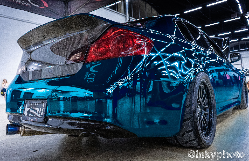 Custom blue chrome wrapped Infiniti G37 sedan with a large carbon fiber wing and aftermarket wheels at an indoor car show.