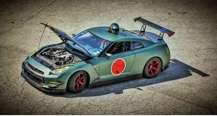 A green Nissan GT-R with a Japanese flag decal, open hood, large rear wing, and red wheels parked on asphalt with a race helmet on the roof.