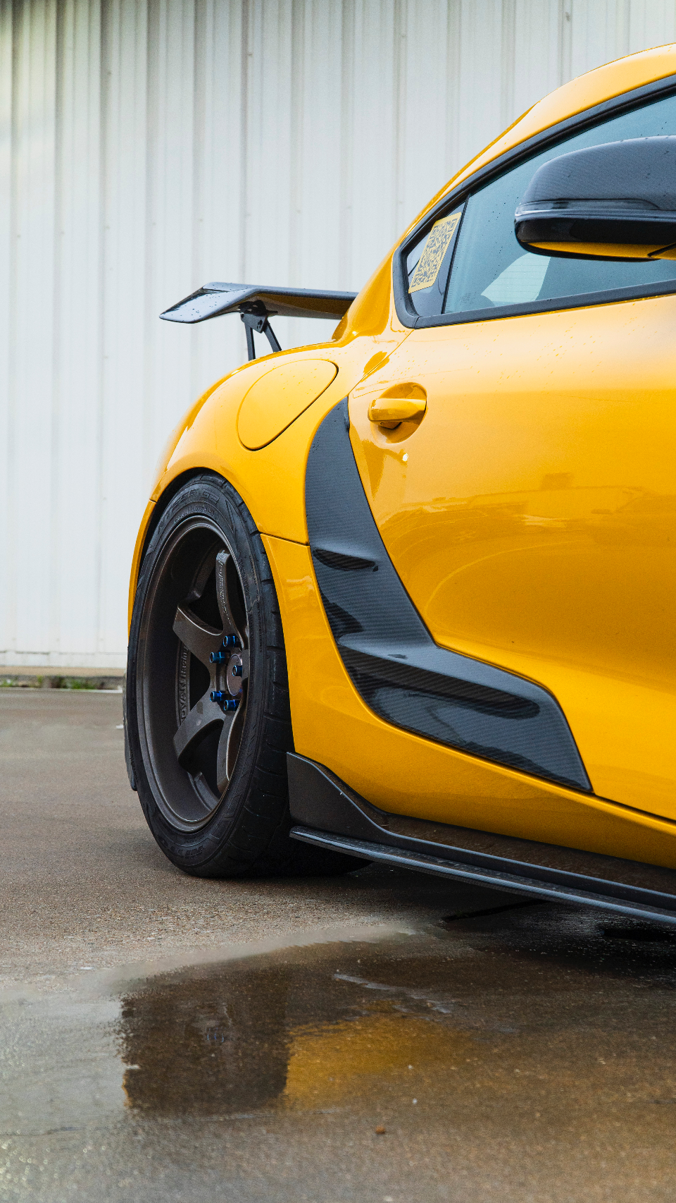 A close-up detail shot of a yellow sports car featuring carbon fiber aerodynamic elements, aftermarket wheels, and a rear wing on wet asphalt.