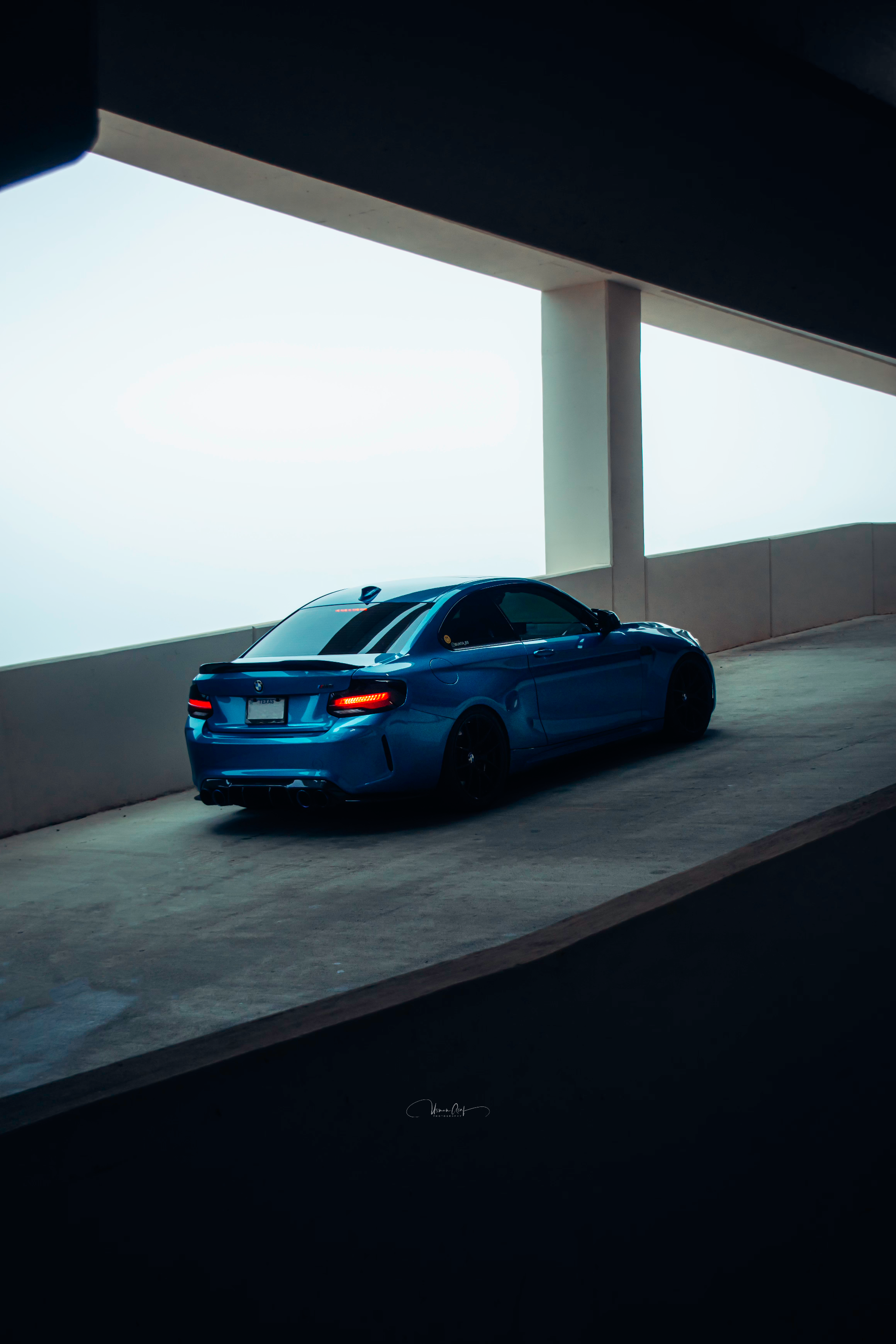 Rear view of a blue BMW M2 with aftermarket spoiler and diffuser in a parking garage.