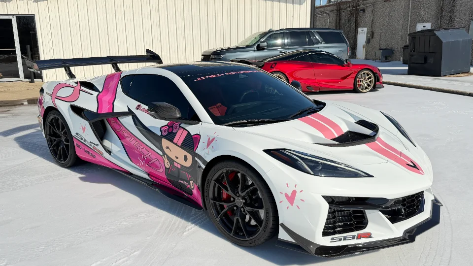 A white Corvette C8 Z06 with a pink and black custom anime-style livery and a large rear wing, parked outdoors.