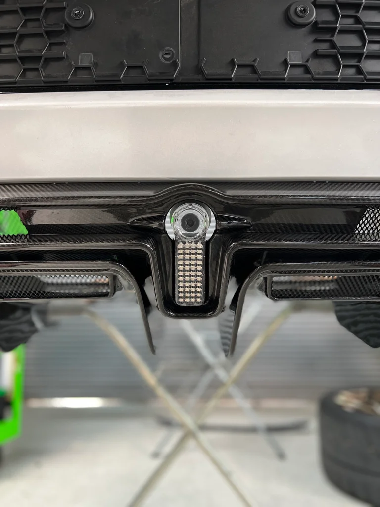 Close-up detail of a car's rear bumper featuring a carbon fiber diffuser, reverse camera, and integrated LED light.