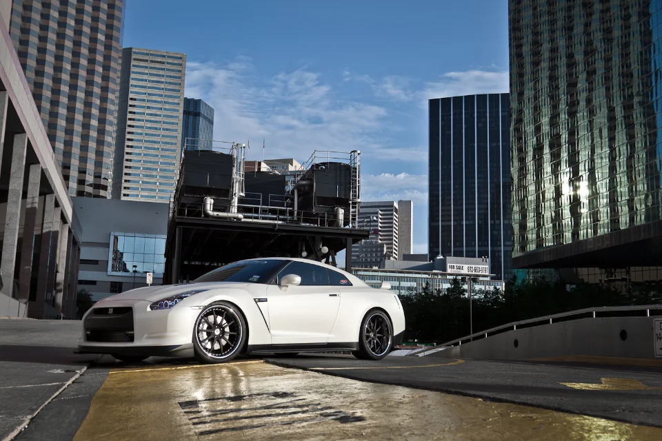 A white Nissan GT-R R35 with aftermarket wheels parked in a city environment with skyscrapers in the background.