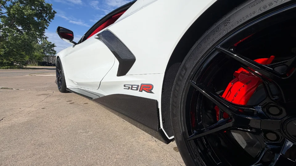 Close-up of a white C8 Corvette's rear wheel, featuring black multi-spoke wheels, red brake calipers, and carbon fiber side skirts.