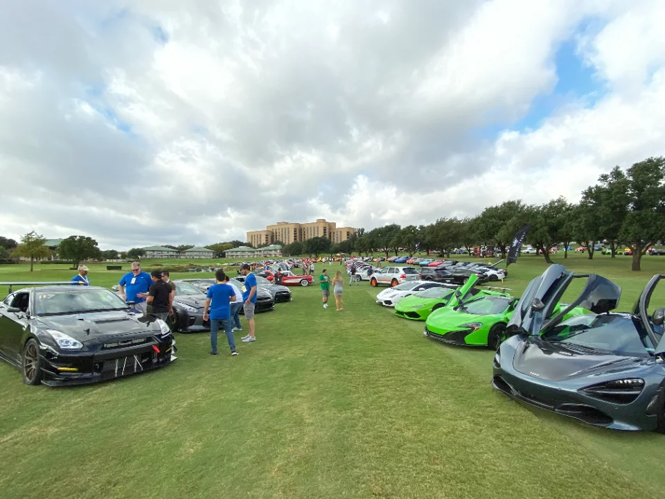 A large outdoor car show with many supercars including a black Nissan GT-R and several McLarens parked on a grassy field with people walking around.