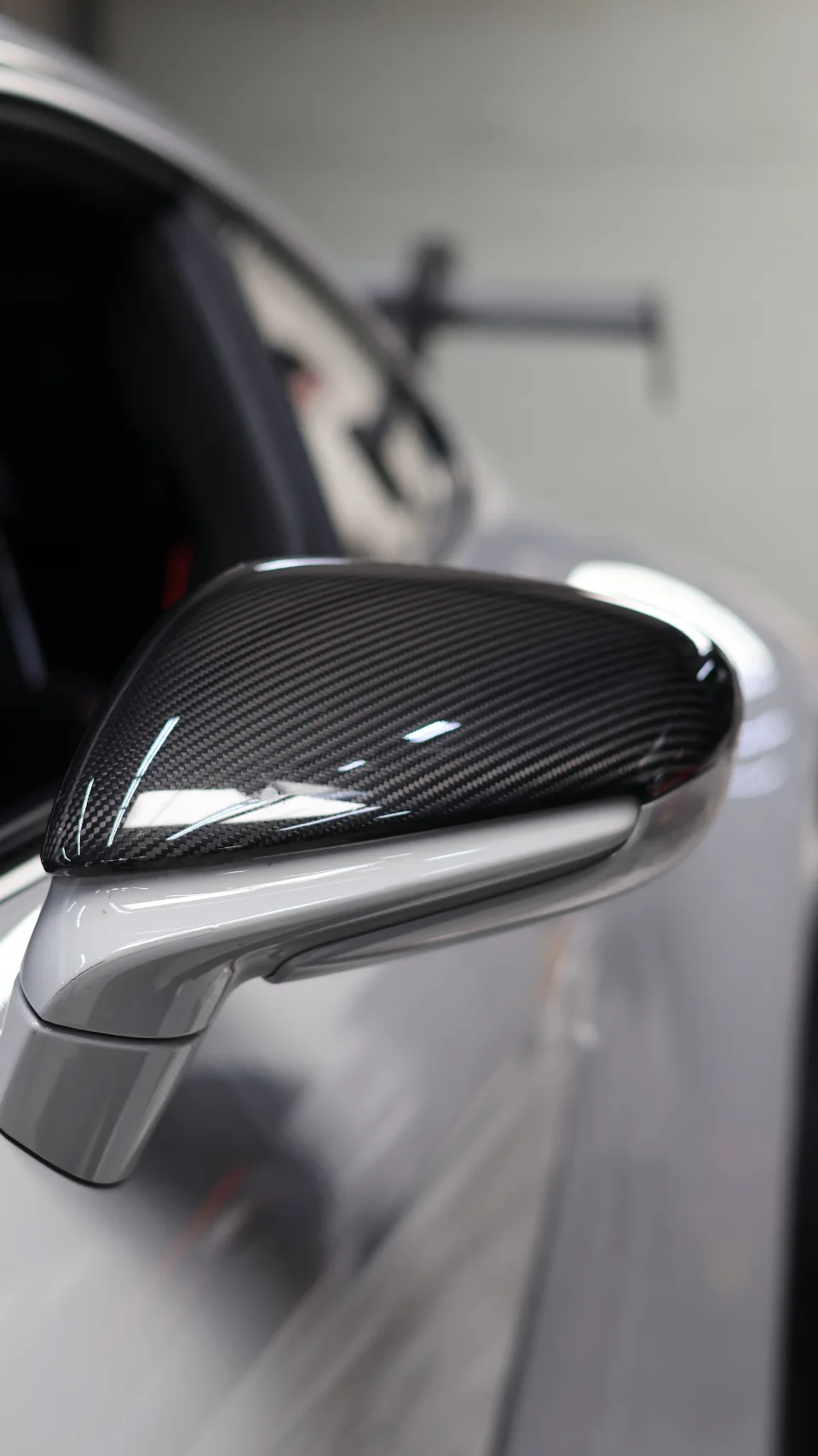 Close-up of a car's carbon fiber side mirror, showcasing the weave pattern and glossy finish on a silver car.