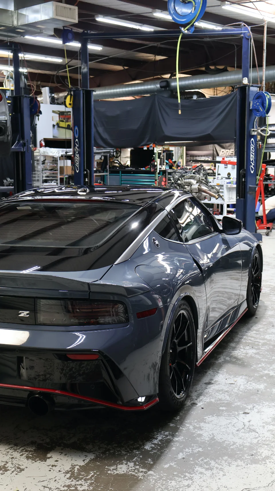 A grey Nissan Z sports car parked in a performance tuning shop with car lifts and tools in the background.