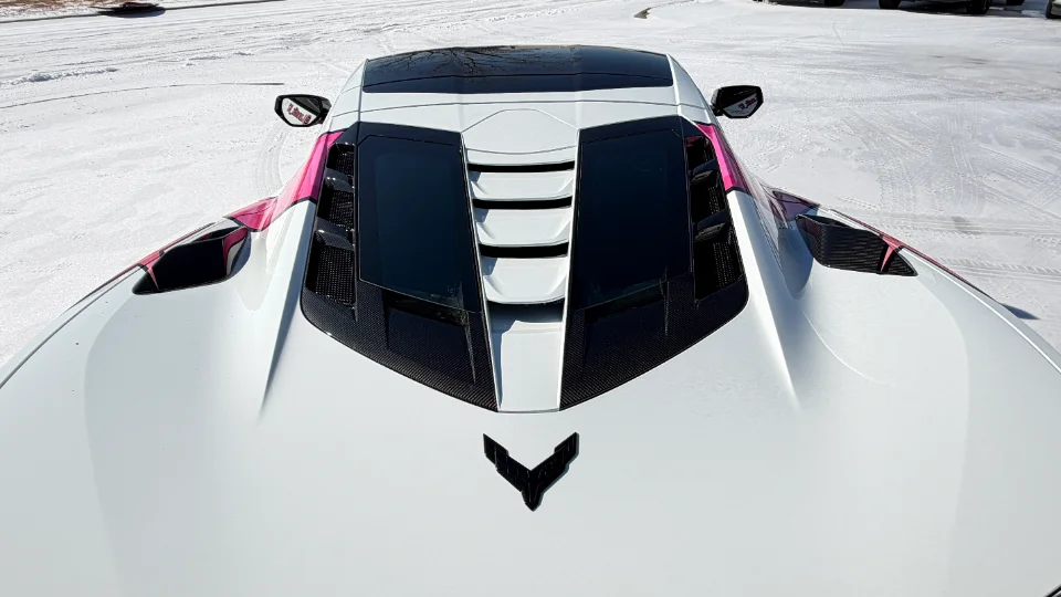 A white Corvette C8 with pink accents and carbon fiber engine cover and vents, parked in a snowy landscape.