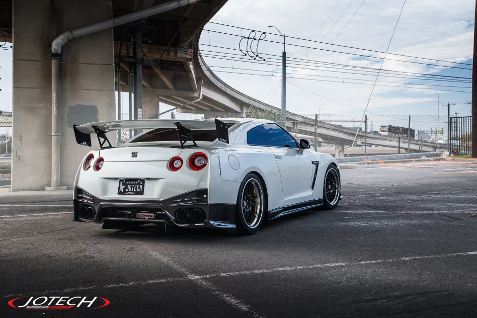 A white Nissan GT-R with a large rear wing and aftermarket wheels parked under a concrete overpass.