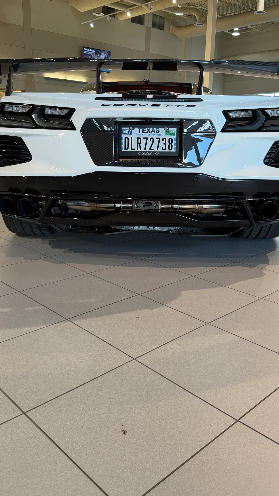Rear view of a white C8 Corvette with a large carbon fiber wing and aftermarket exhaust system in a showroom.