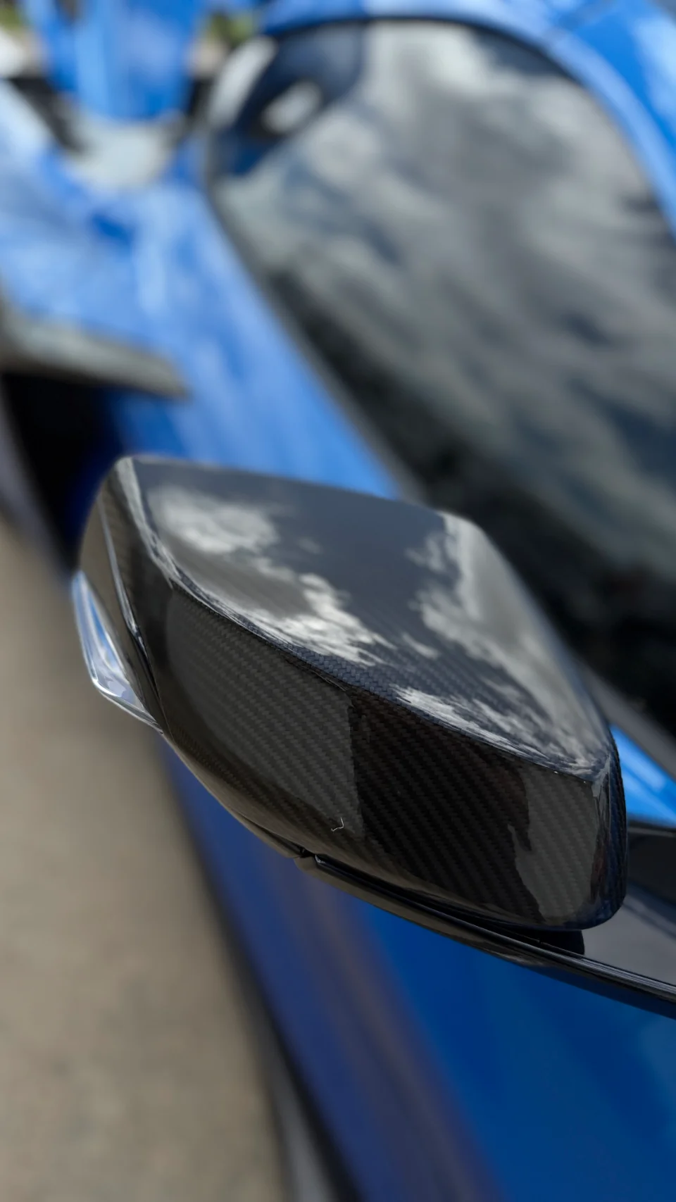 A close-up of a blue car's carbon fiber side mirror, reflecting clouds in its glossy surface.