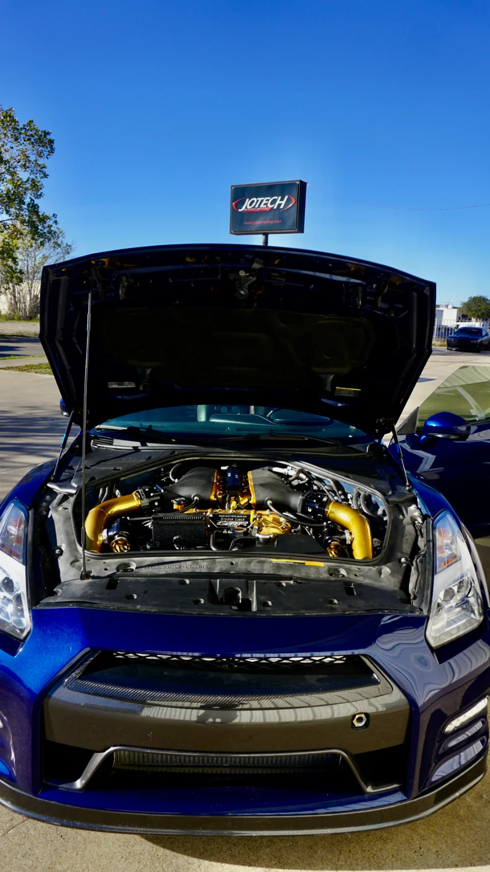Blue Nissan GT-R with its hood open, showcasing a custom gold engine bay at Jotech Motorsports.