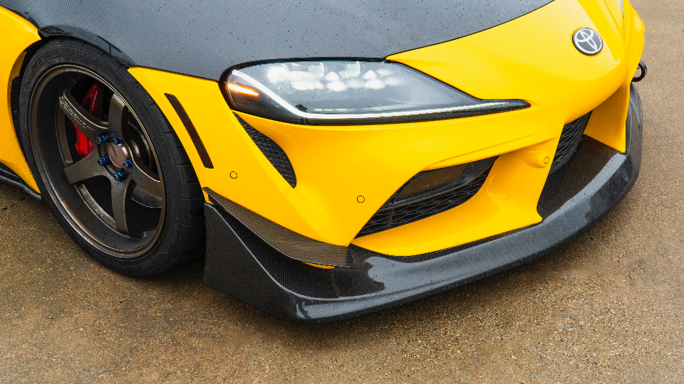 Close-up of a yellow Toyota Supra's front end, showcasing carbon fiber aero, aftermarket bronze wheels, and LED headlights.