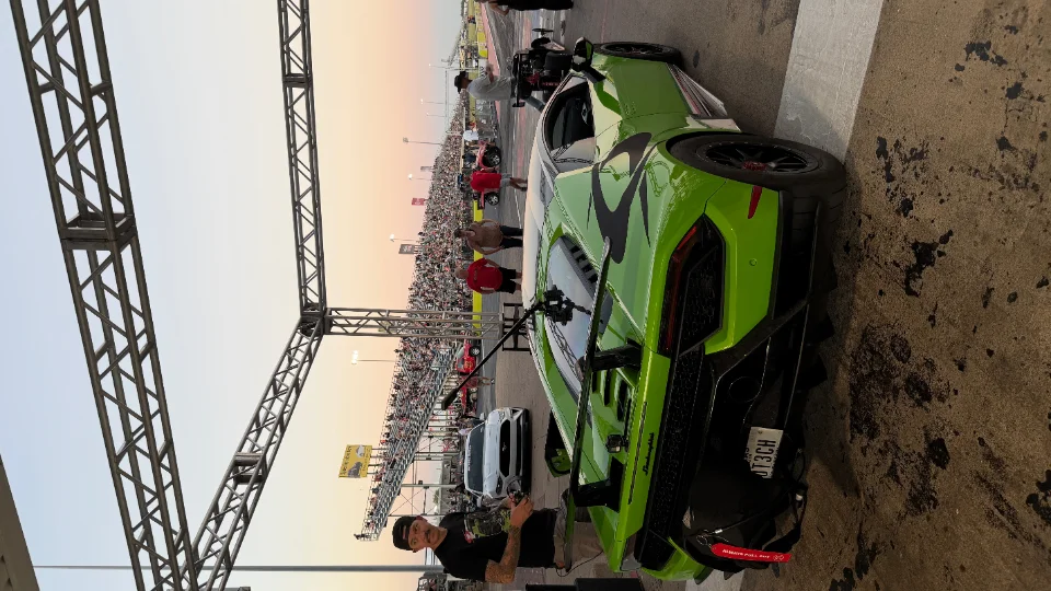 A bright green Lamborghini Huracan with black accents parked at a drag strip, with a crowd of spectators in the background at dusk.
