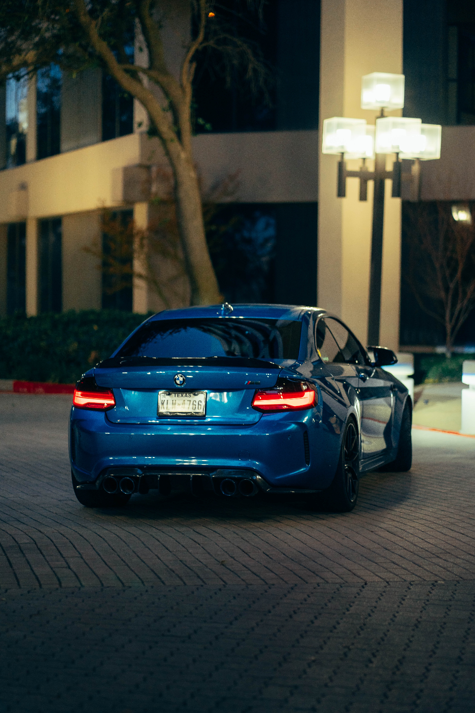 Rear view of a blue BMW M2 with quad exhaust and a diffuser, parked at night under streetlights.