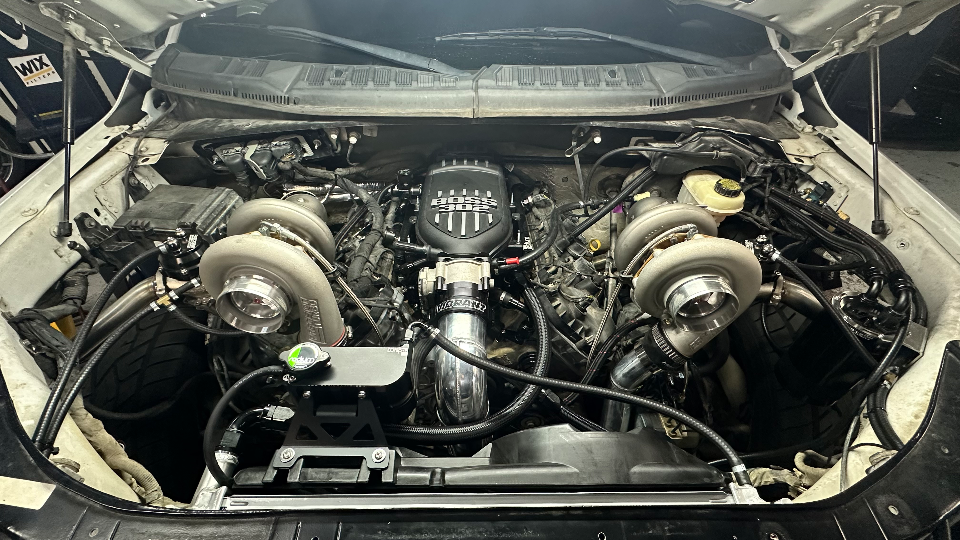 A detailed view of a twin-turbocharged Ford F-150 engine bay featuring a BOSS 302 intake manifold and custom piping.