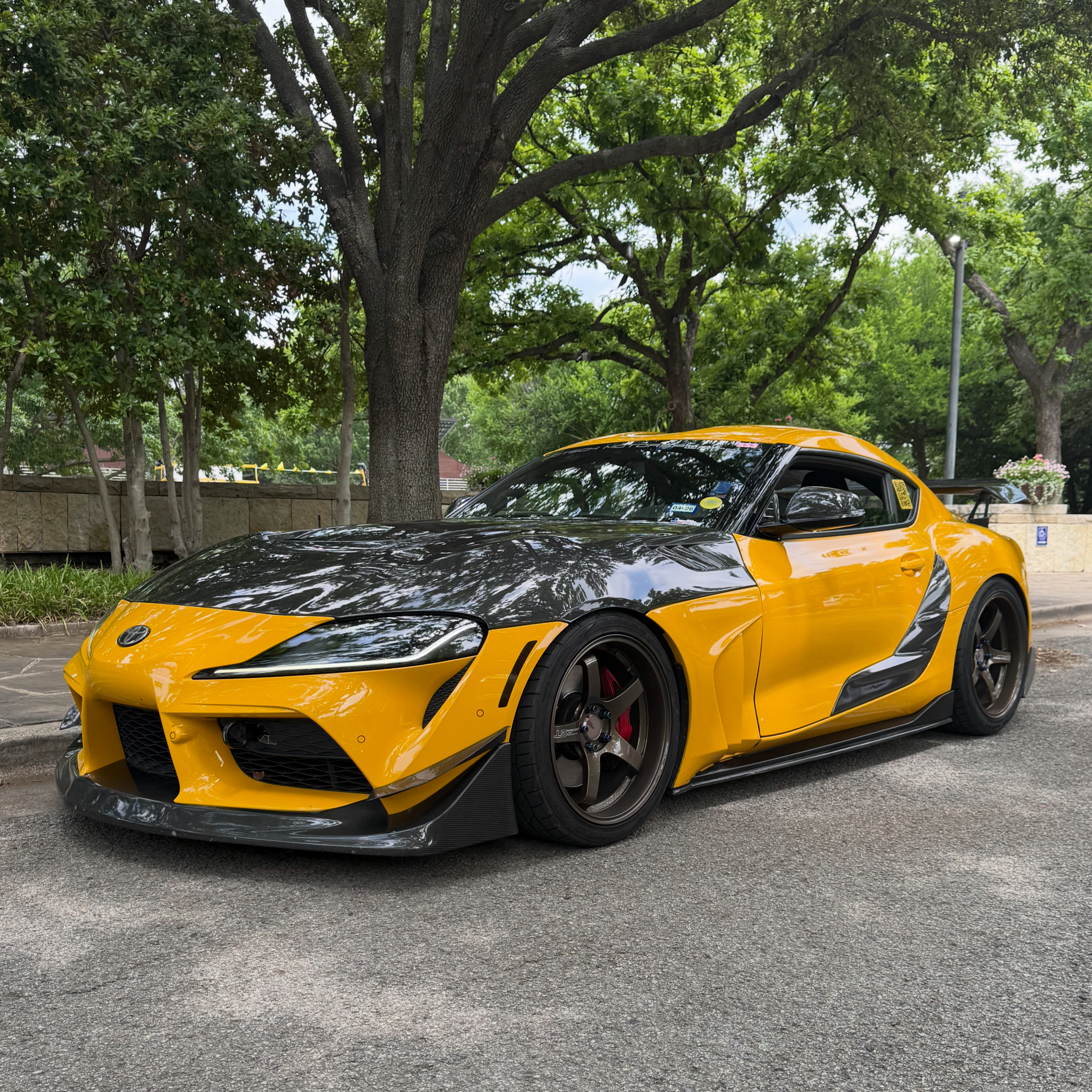 A vibrant yellow Toyota Supra with a carbon fiber hood, front splitter, side skirts, and aftermarket bronze wheels parked on a street.