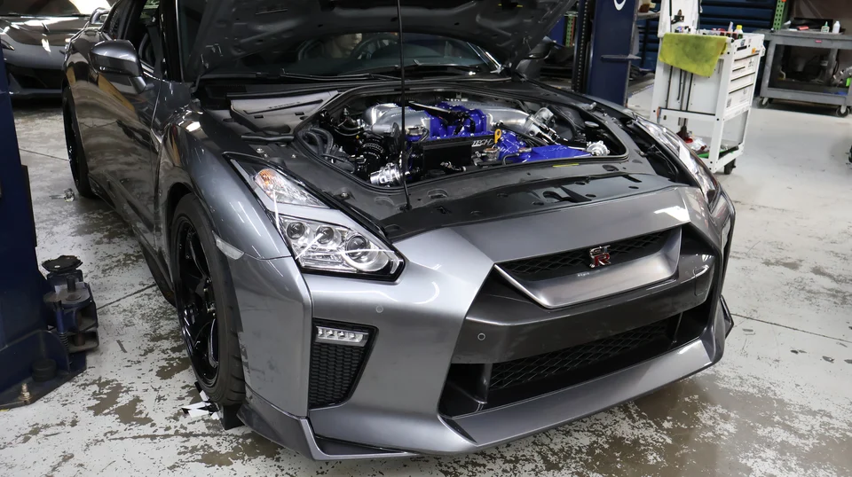 A grey Nissan GT-R with its hood open, showcasing a modified engine bay with blue and silver performance parts in a shop.
