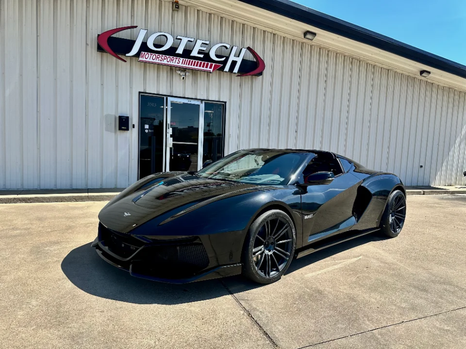 A sleek black custom supercar parked in front of the Jotech Motorsports shop on a sunny day.