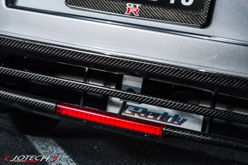 Close-up of a Nissan GT-R's rear bumper, showcasing carbon fiber aerodynamic elements and a GReddy performance cooler.