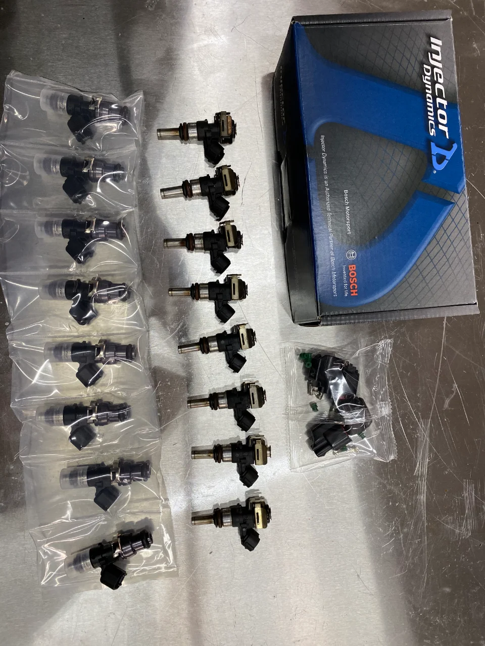 New Injector Dynamics fuel injectors in their packaging, along with loose injectors, displayed on a metal workbench.