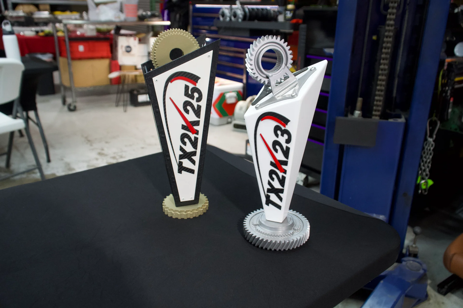 Two TX2K event trophies, one for 2023 and one for 2025, displayed on a table in a shop.