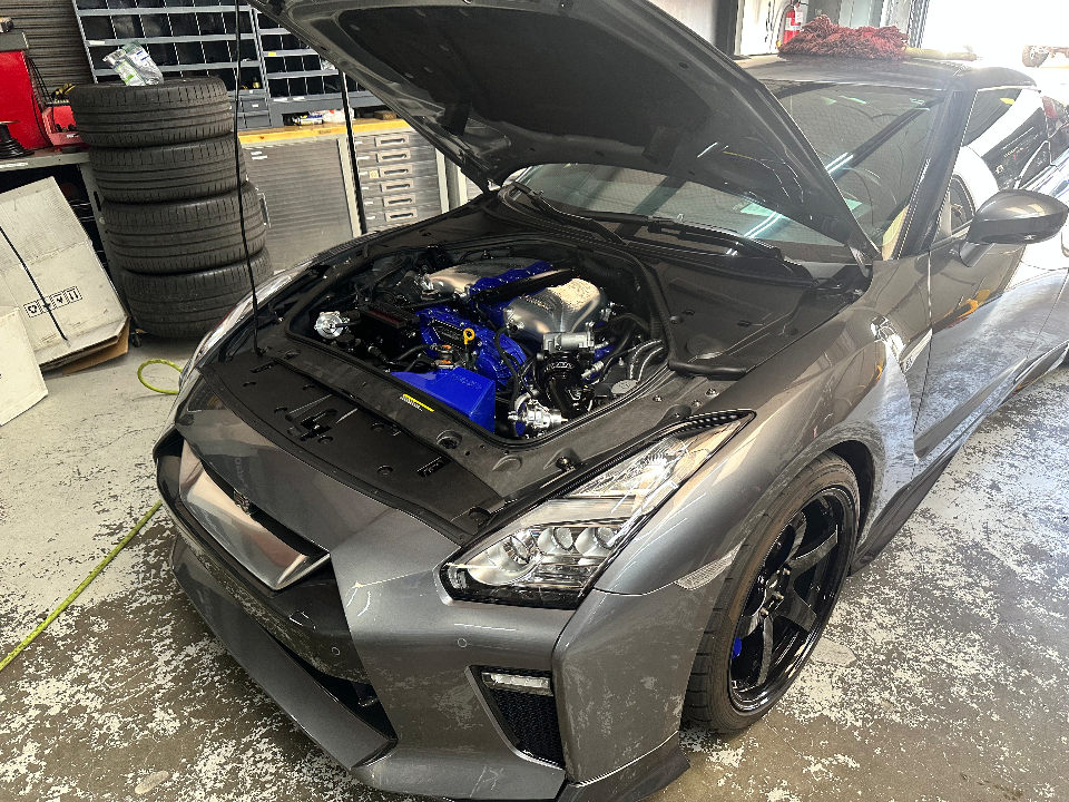A grey Nissan GT-R with its hood open, showcasing a custom blue engine bay in a performance shop.