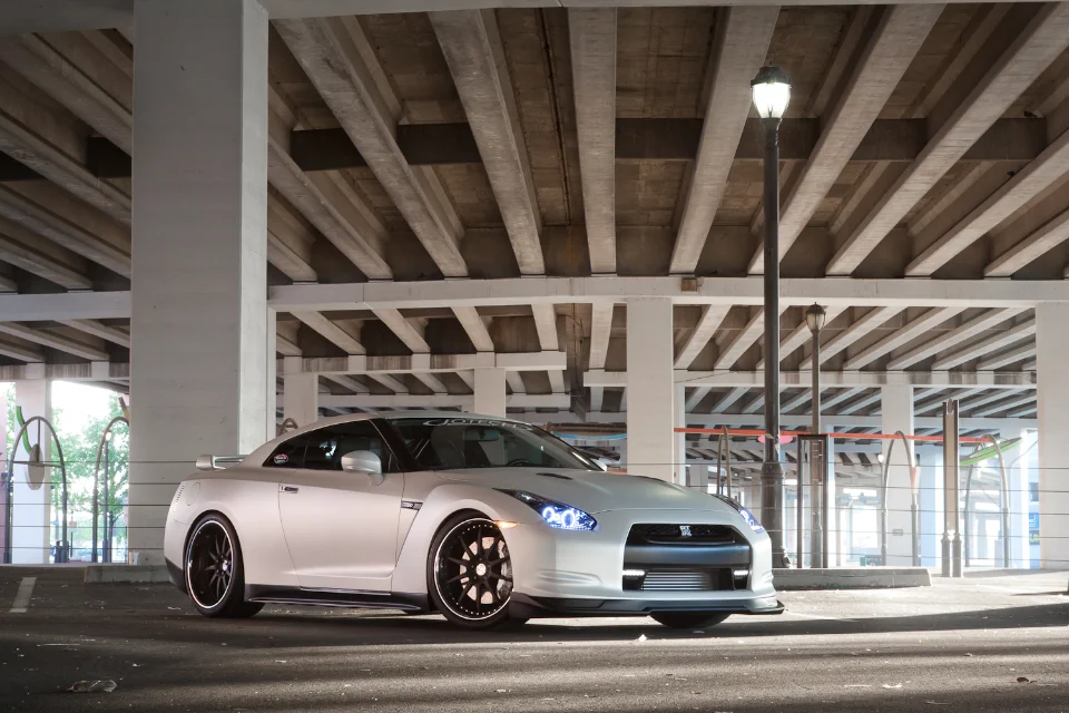 A white Nissan GT-R R35 with custom black wheels parked under a concrete overpass with a street lamp.