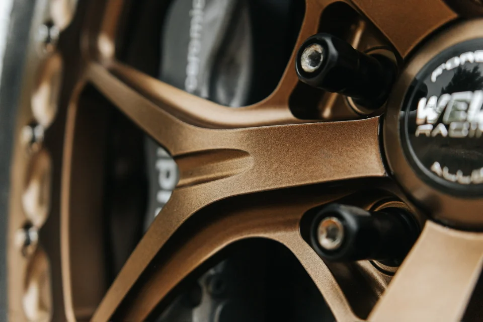 A close-up detail shot of a bronze Weld Racing wheel with black lug nuts and a Porsche ceramic brake caliper.