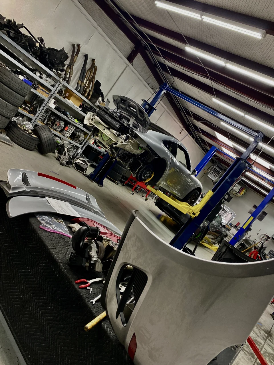 A silver Porsche 911 on a lift in a performance tuning shop, with various car parts and tools on shelves and tables.