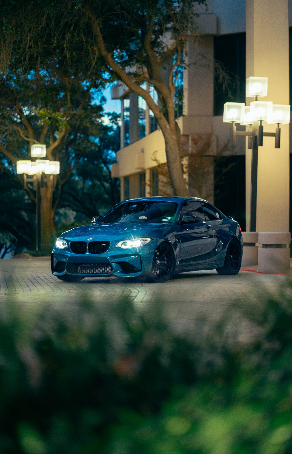A vibrant blue BMW M2 coupe parked at night in front of a modern building, illuminated by street lights.
