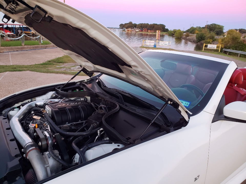 White Infiniti G37 convertible with a Stillen supercharger visible in the engine bay, parked by a lake at sunset.