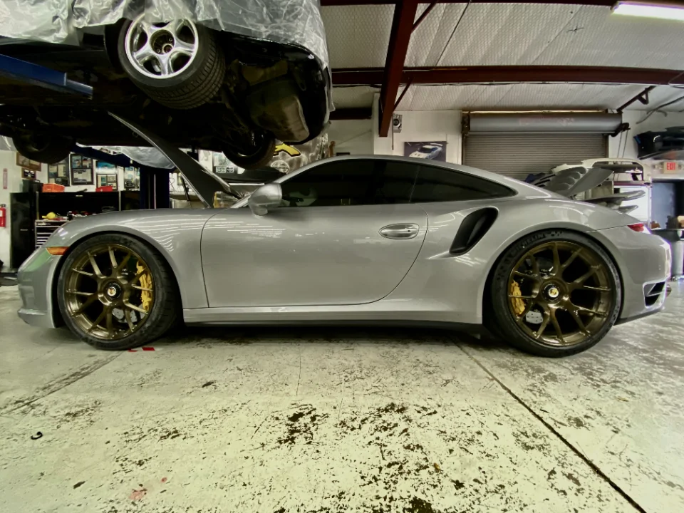 A silver Porsche 911 Turbo S with custom bronze wheels parked in a Jotech Motorsports shop with another car on a lift.
