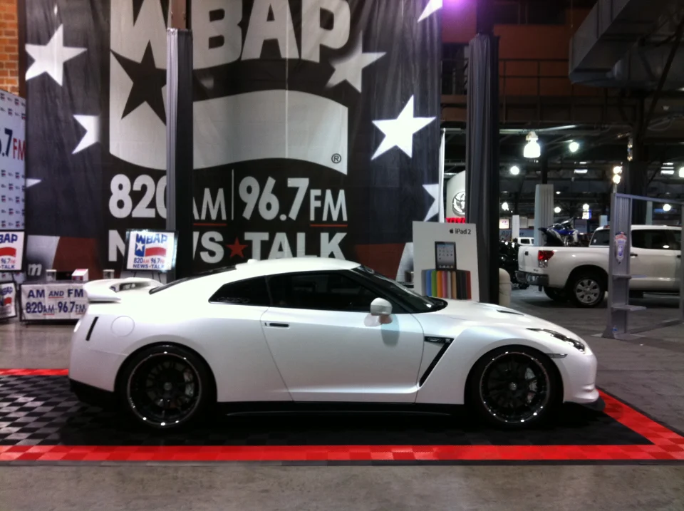 White Nissan GT-R on display at an indoor car show with large WBAP radio banners in the background.