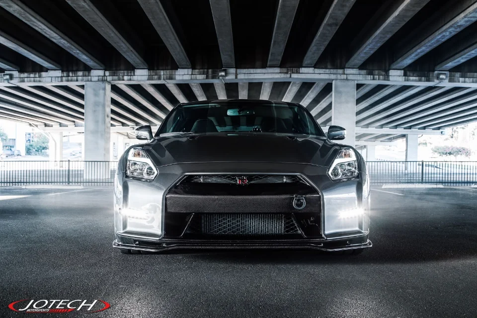 Front view of a grey Nissan GT-R with carbon fiber aero and a visible intercooler, parked under an overpass.
