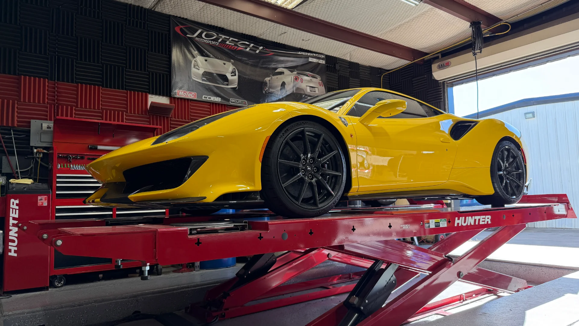 A bright yellow Ferrari-like sports car is elevated on a red Hunter lift inside a Jotech Motorsports performance shop.