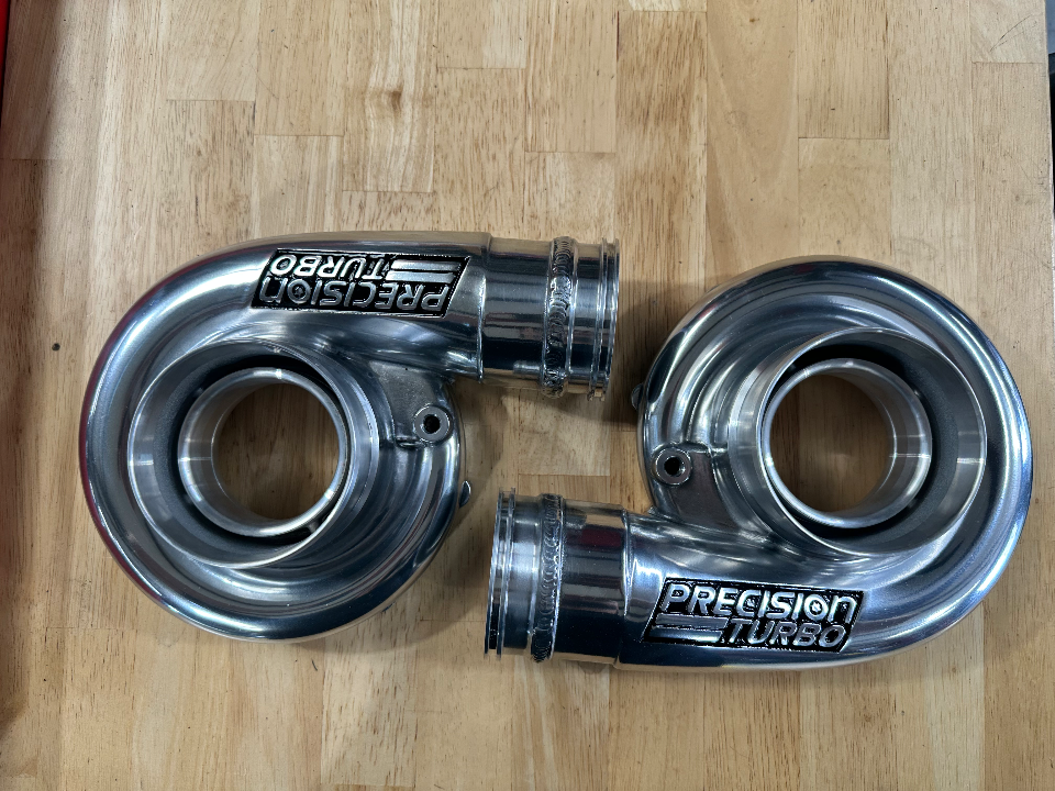 Two shiny Precision Turbo compressor housings on a wooden surface, ready for a high-performance build.