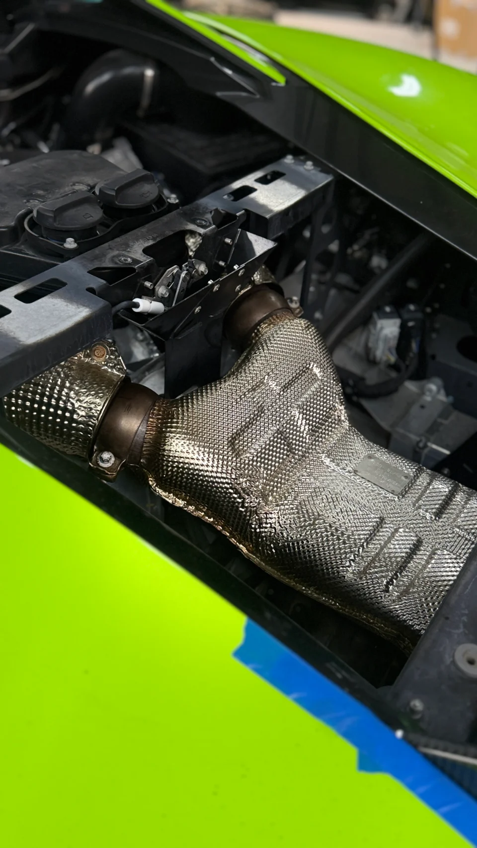 A close-up of an aftermarket exhaust manifold with a heat shield installed in the engine bay of a green Lamborghini.