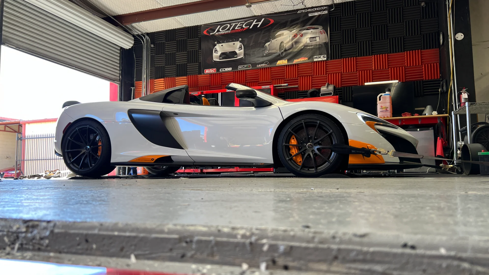 A white McLaren supercar is strapped down on a dynamometer at Jotech Motorsports, ready for performance tuning.