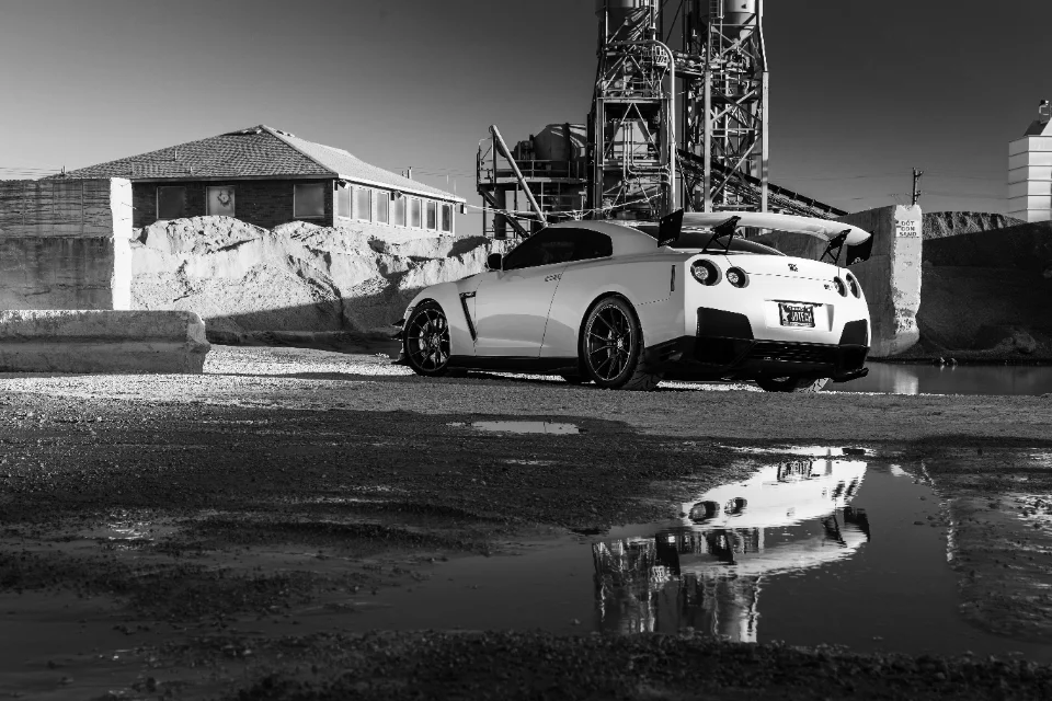 A white Nissan GT-R with an aftermarket wing and wheels parked in an industrial setting with a reflection in a puddle.