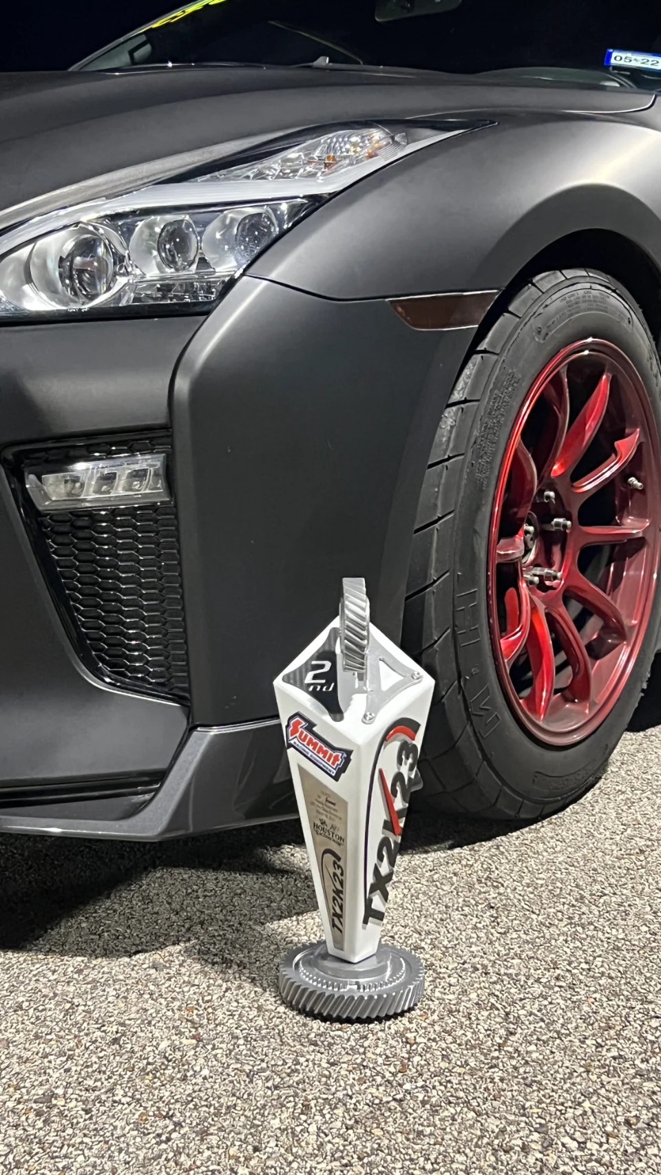 A matte black Nissan GT-R with red wheels parked, a TX2K23 2nd place trophy in front of its headlight.