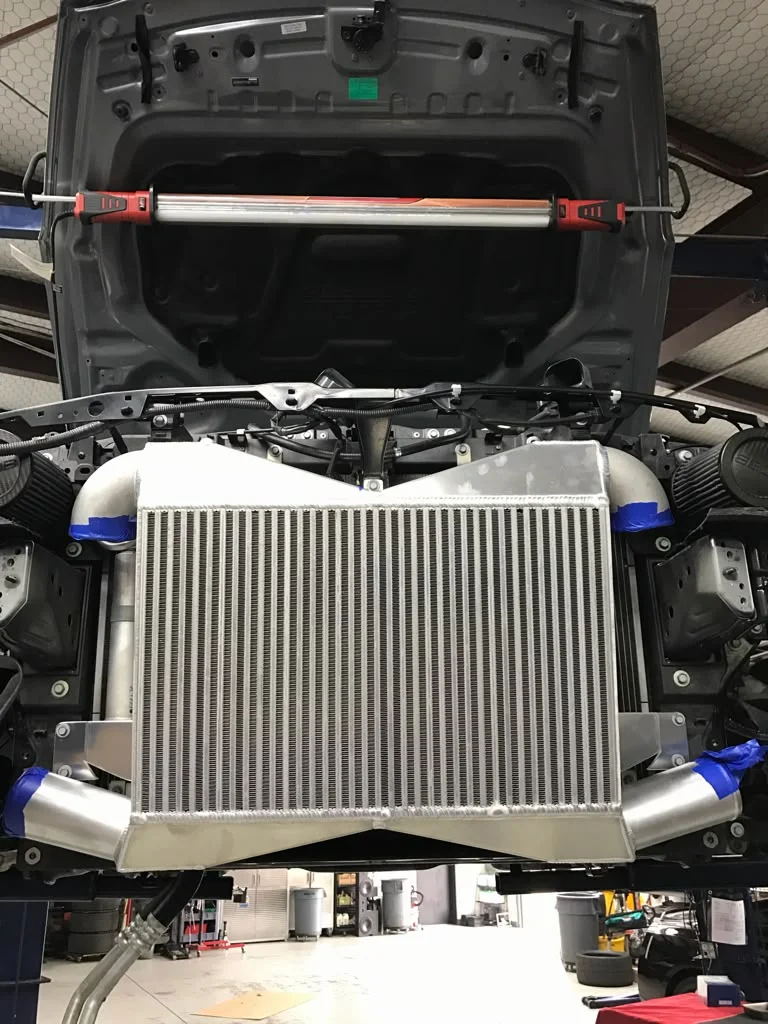 Front view of a car on a lift in a shop, showcasing a large aftermarket intercooler and boost piping installation.