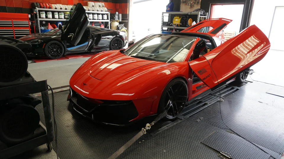 A custom red sports car with its doors open, strapped to a dynamometer in a performance tuning shop, with a black car in the background.