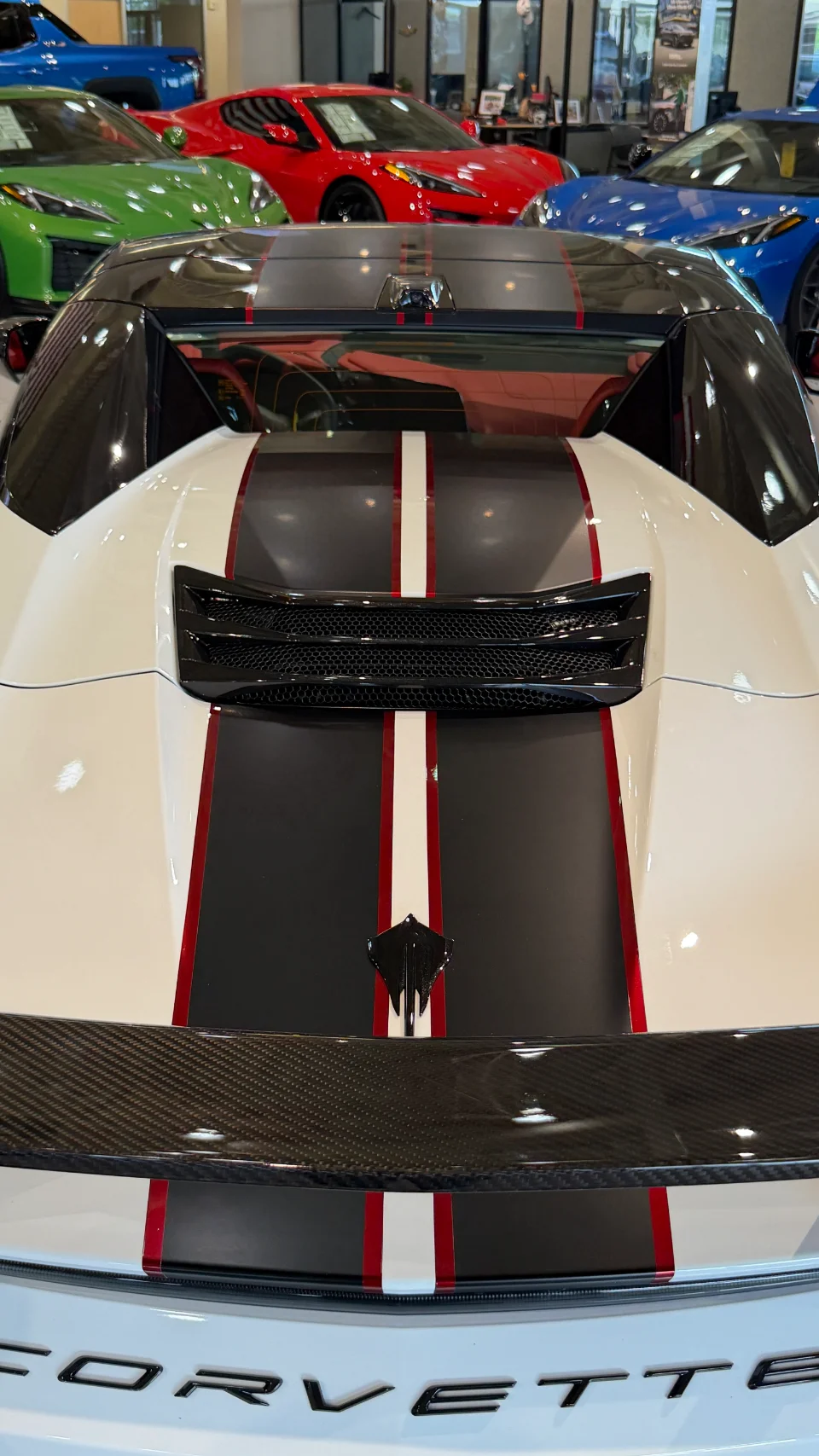 Rear view of a white Corvette C8 with black and red racing stripes and a carbon fiber spoiler in a showroom.