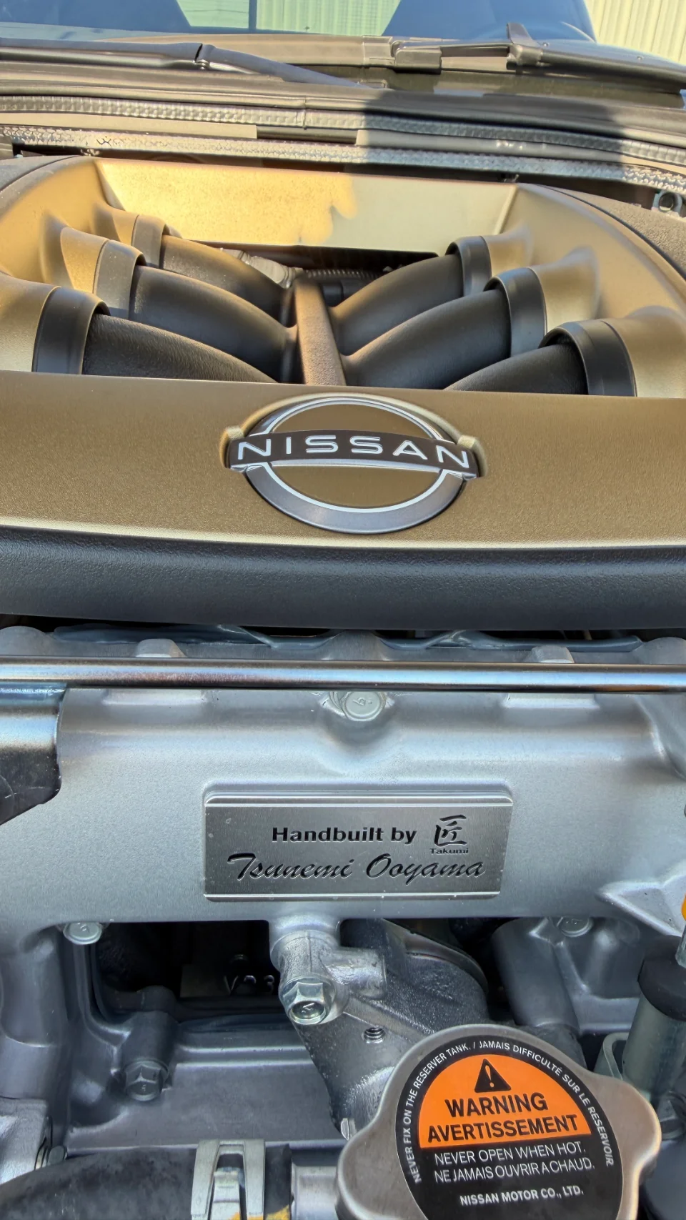 Close-up of a Nissan GT-R VR38DETT engine bay with a gold engine cover and a 'Handbuilt by Tsuneaki Ooyama' plaque.