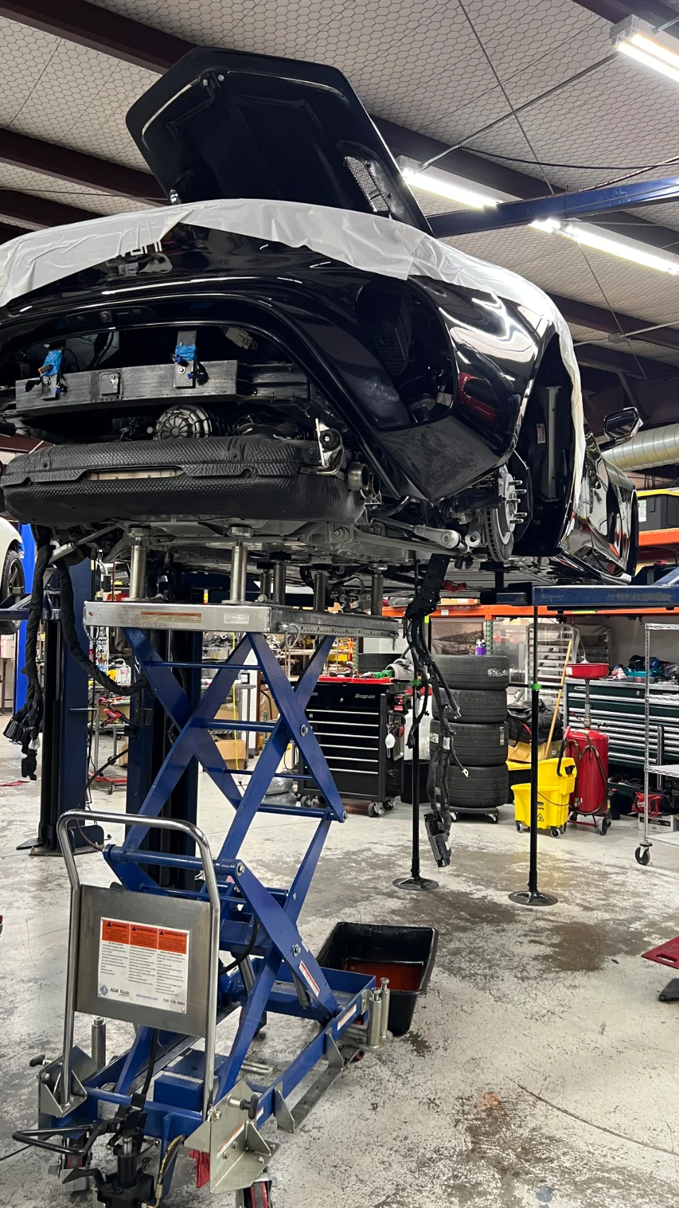 A black performance car elevated on a lift in a mechanic shop, with its rear open and exhaust system visible during maintenance.