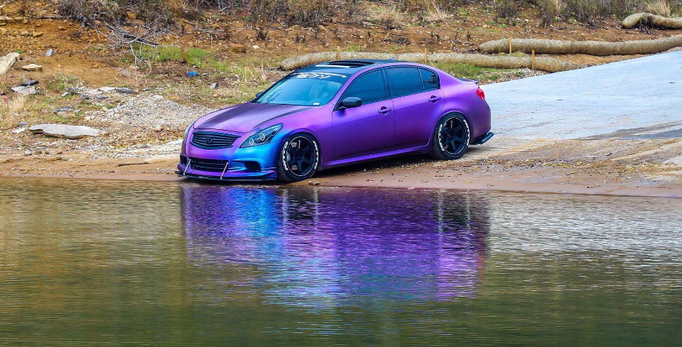 A custom purple and blue color-shifting Infiniti G37 sedan parked at the edge of a lake with its reflection in the water.