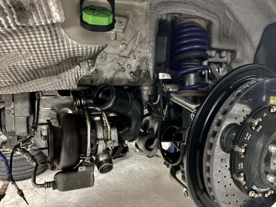 Close-up view of a car's undercarriage showing a turbocharger, blue coilover suspension, and large brake rotor during a shop service.