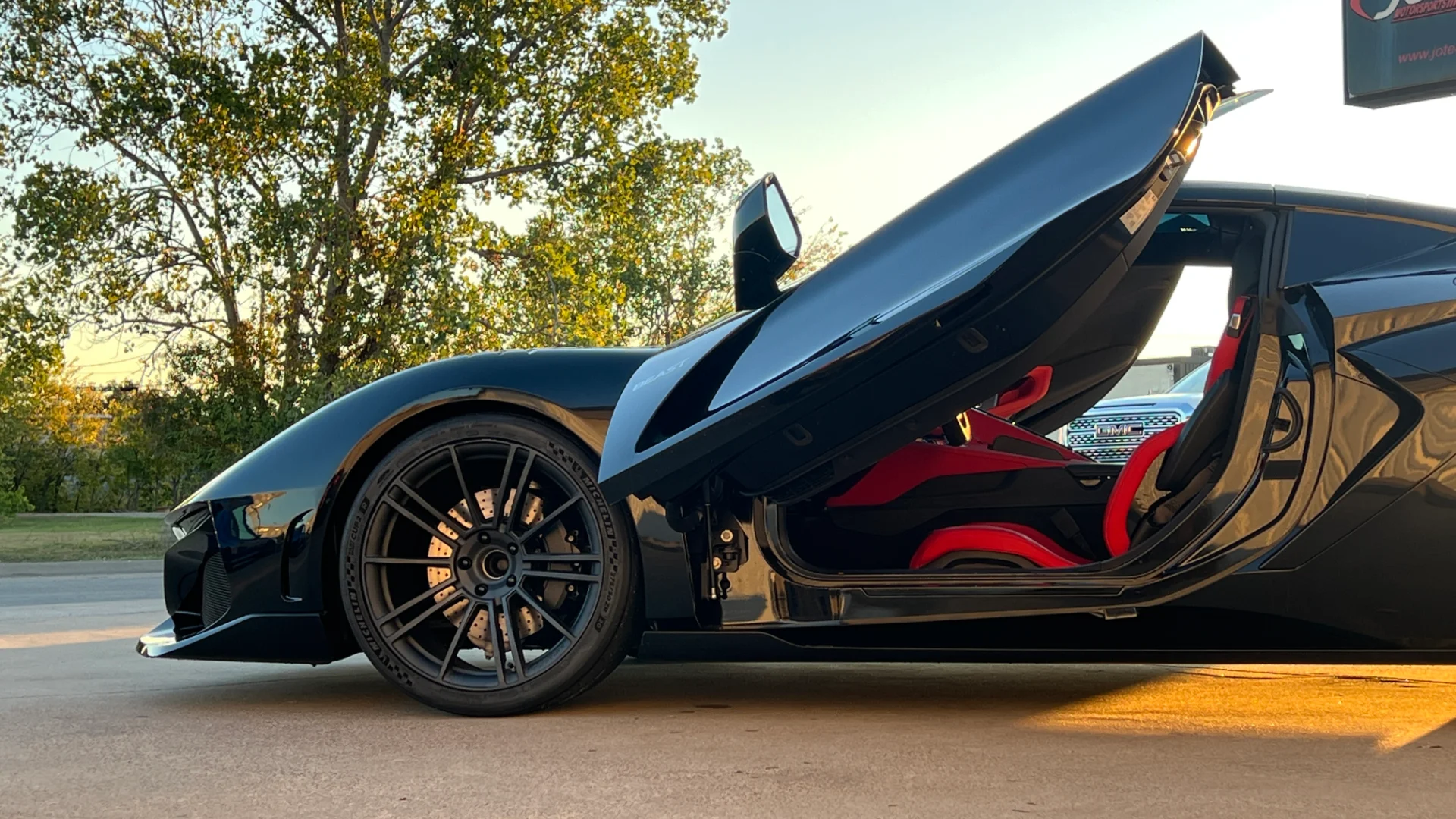 A black exotic sports car with unique upward-opening doors and a striking red interior, parked outdoors near a Jotech Motorsports sign.