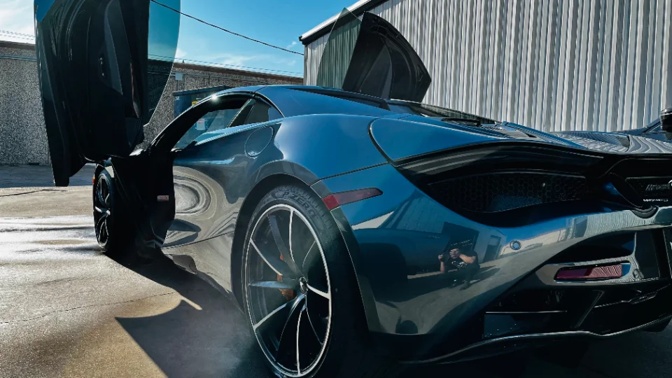 A dark grey McLaren supercar with its butterfly doors open, parked outside a building on a sunny day.